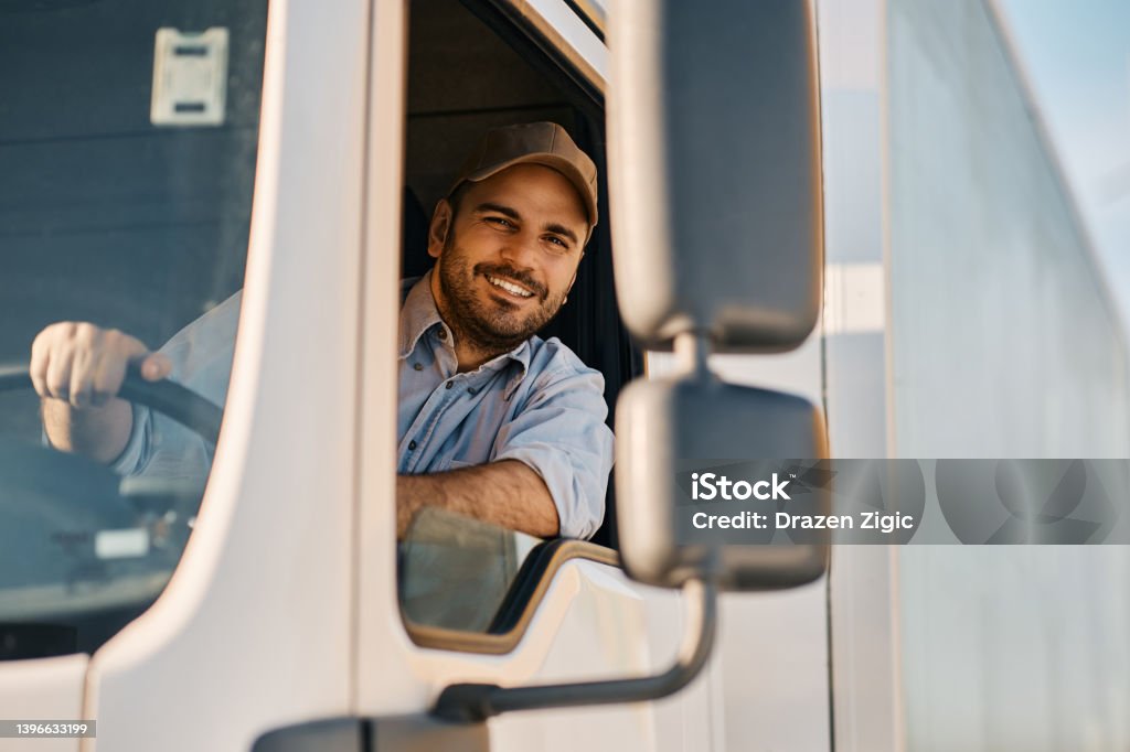 Happy truck driver looking through side window while driving his truck. Happy professional truck driver driving his truck and looking at camera. Copy space. Truck Driver Stock Photo