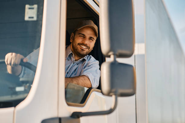 Happy truck driver looking through side window while driving his truck. Happy professional truck driver driving his truck and looking at camera. Copy space. truck stock pictures, royalty-free photos & images
