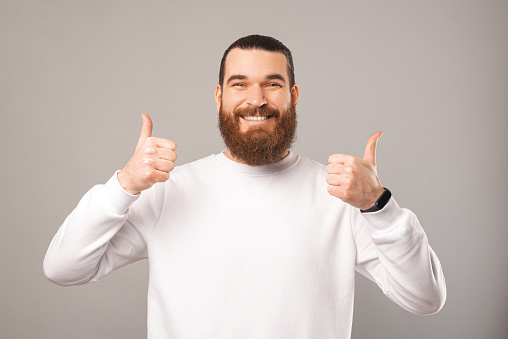 Friendly bearded man wearing white is showing two thumbs up while smiling at the camera. Studio shot over grey background.