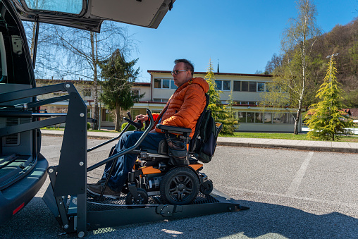 Disabled man in a motorized wheelchair when entering a van on an electric lift. In front of the vehicle, on the vehicle.