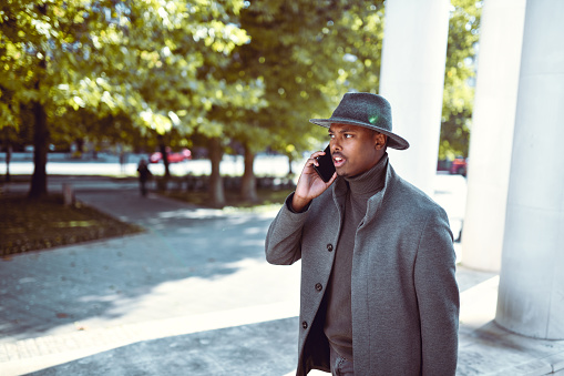 Modern African Male In Coat Having Phone Call Conversation While Walking In A Park