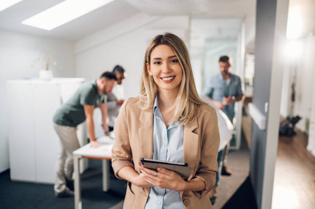 Businesswoman posing and smiling during a meeting in an office Smiling confident business leader looking at camera and standing in an office at team meeting. Portrait of confident businesswoman with colleagues in boardroom. Posing while holding digital tablet. girl work stock pictures, royalty-free photos & images