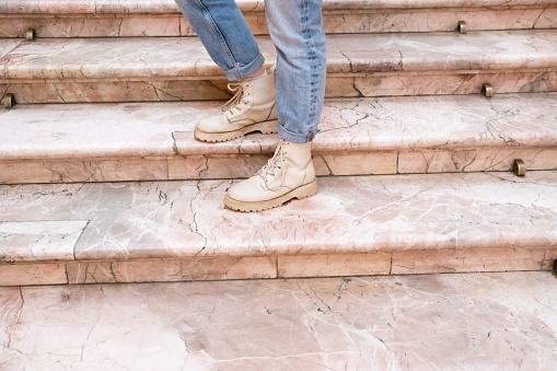 Woman's legs in blue twisted jeans and in beige boots on pink marble staircase. Front view, copy space.