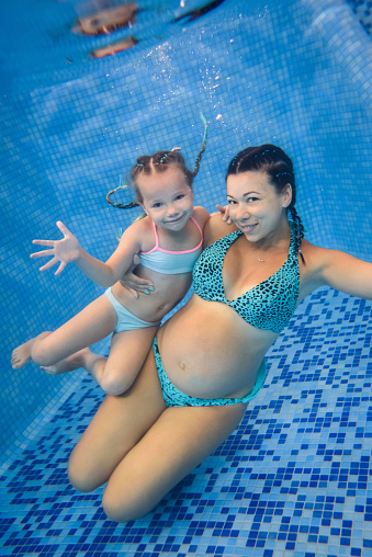 Mother and daughter learn to swim on swimming lesson, doing physical exercises, dive underwater with in pool. Healthy lifestyle, active parents. Pregnant woman swims underwater.