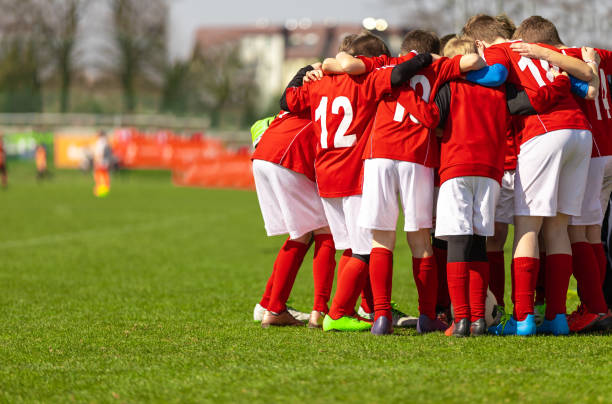 kinderfußballmannschaft mit trainer in der gruppe vor dem spiel. kinder im grundschulalter hören gemeinsam zu, um motivierende sprache zu coachen. jungen in roten fußballtrikots - mannschaftsfußball stock-fotos und bilder