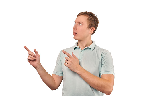 Surprised young guy with funny face in grey T-shirt pointing to left, white isolated background. Shocked amazed face of young man in casual style. Portrait of excited man gaping mouth in surprise