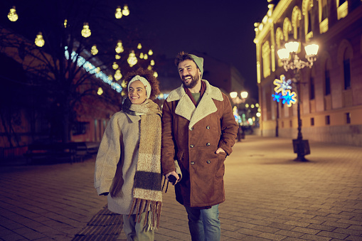 Young happy couple holding hands while walking during Christmas night on the city street.