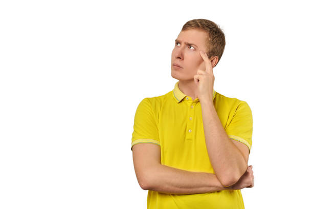 Thoughtful guy in yellow T-shirt looking to left isolated on white background. Pensive young man thinking, guy putting hand to his head and looking to left, philosophical reflection concept