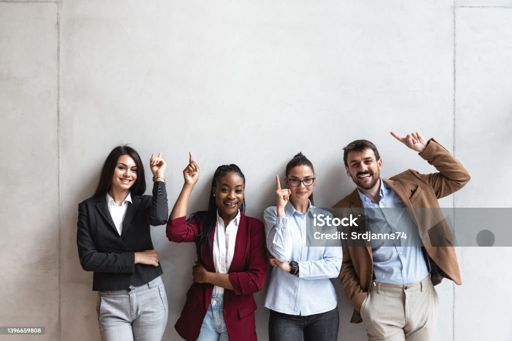 Group of young happy successful business team people pointing finger up and smiling. Freelancers or exchange students showing above and smile looking at the camera. Meeting Stock Photo