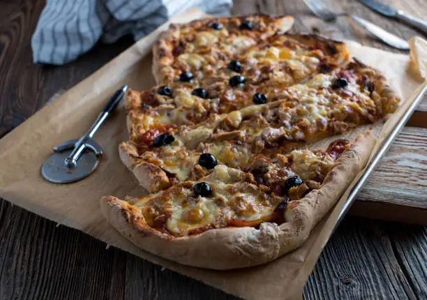 Fresh baked pizza with tuna, tomatoes, onions, black olives and mozzarella cheese. Served with pizza cutter on a baking tray on wooden table background.
