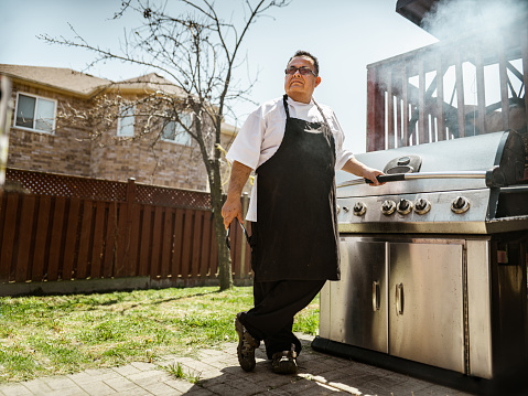 Mature Latin Chef of Mexican background posing by the backyard BBQ. He is wearing eyeglasses, chefs robe with apron. Interior of private home  kitchen.