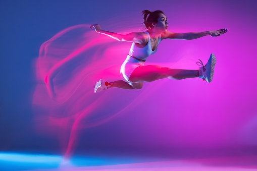 Long jump. Young athlete, runner training isolated on blue studio background in mixed pink neon light. Sportive girl in white sportswear practicing in run or jogging. Healthy lifestyle concept.