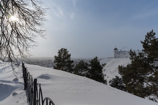 The city of Tobolsk (Siberia, Russia) in the haze from the morning frost. View from a high snow-covered hill with historic buildings of the Kremlin and tall pines through which the sun breaks