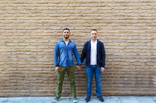 Gay couple holding hands posing in front of a brick wall. Homosexual relationship concept.