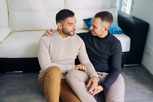 Gay couple sitting near the couch at home in a romantic moment. Homosexual relationship concept.