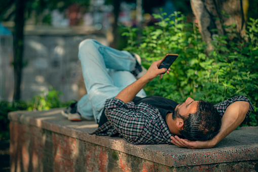 In this day image, an Asian/Indian young man uses a smartphone lying on his back under the tree shade in a public park. There are lush foliages of plants in the background.