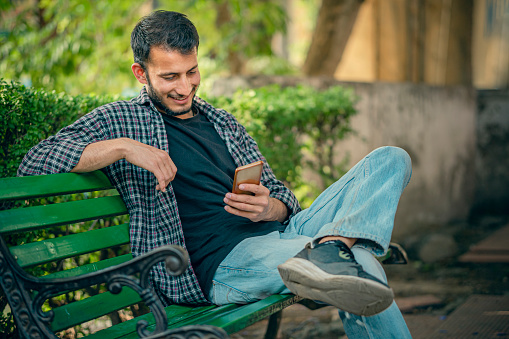 In this day image, an Asian/Indian young man uses a smartphone sitting on the park bench under the tree shade in a public park. There are lush foliages of plants in the background.