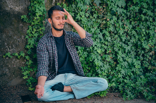 In this day image with copy space, an Asian/Indian young man sits, does breathing exercises (Anuloma Viloma Pranayama), and meditation in a public park under the tree shade.  There are lush foliages of creeper plants in the background.