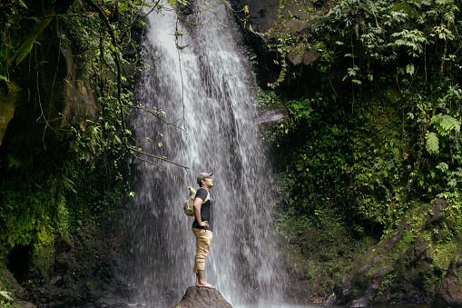 asian man walking  across a rocky surface to get to a waterfall in the forest