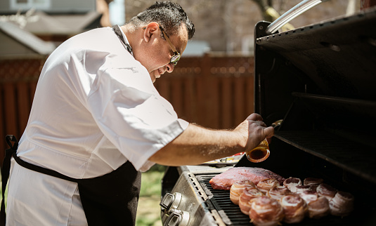Mature Latin Chef grilling meat on BBQ in the backyard. He is wearing chefs robe with apron. Exterior of private home backyard.