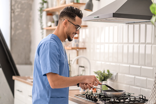Smiling man in eyeglasses standing at the gas stove and stirring food in the frying pan