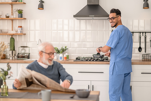 Smiling caregiver stirring food in the frying pan and looking at a pensioner pointing at something in the newspaper article