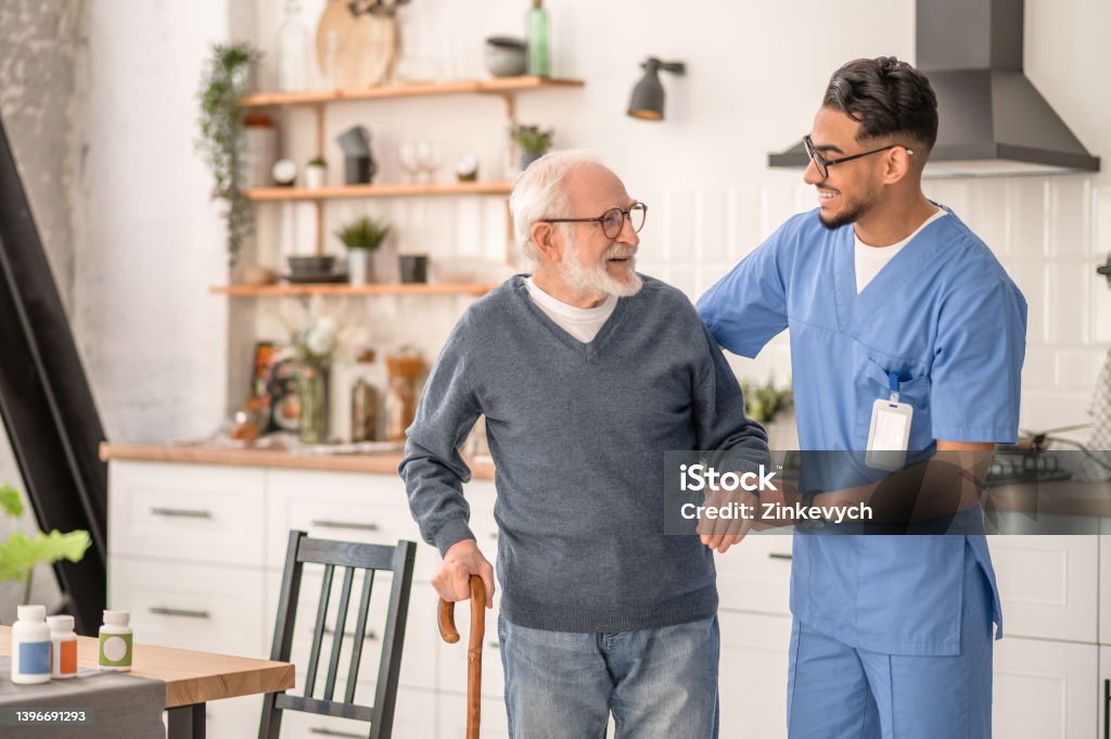 Medical worker helping his patient to move around the apartment Smiling friendly in-home male nurse in uniform supporting an old man with a walking stick Home Caregiver Stock Photo