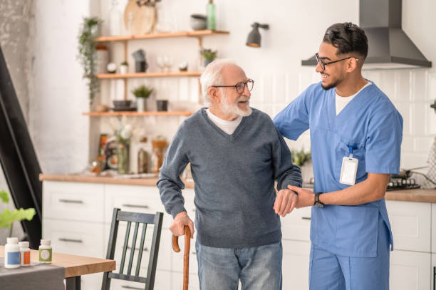 Medical worker helping his patient to move around the apartment Smiling friendly in-home male nurse in uniform supporting an old man with a walking stick home caregiver stock pictures, royalty-free photos & images