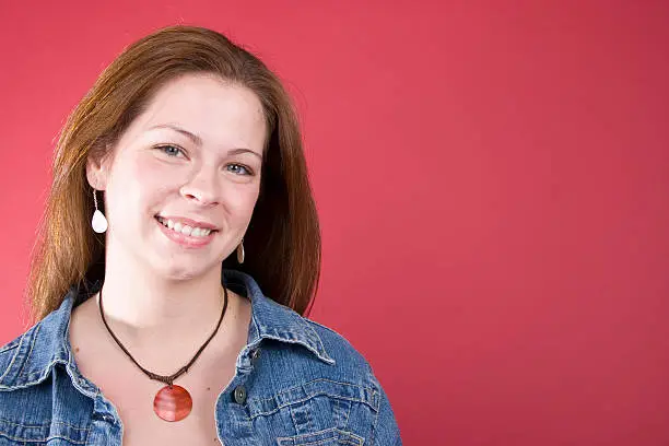 A smiling young adult female in a jean jacket on a red background.