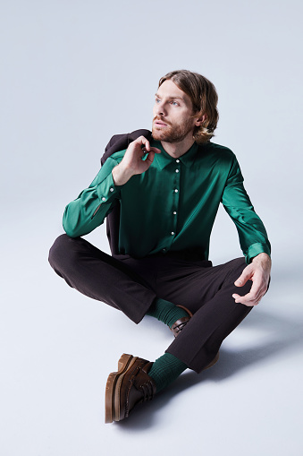 Trend portrait of male fashion model posing casually wearing suit with teal green silk shirt and sitting on floor