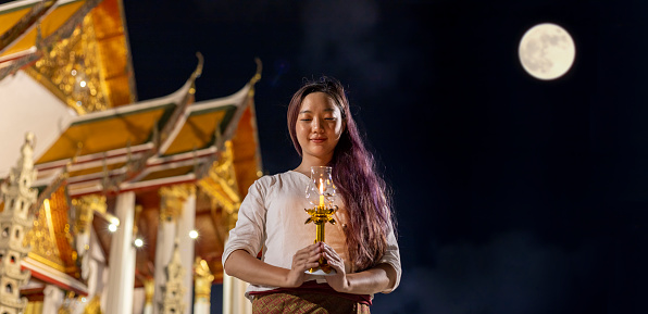 Asian buddhist woman lighting up the candle to pay homage to Lord Buddha on Vesak day at the full moon night inside golden temple for faith, belief and traditional religion culture