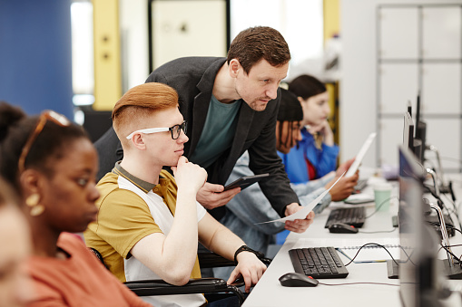 Vibrant side view portrait of male teacher helping student using computer in college classroom, copy space