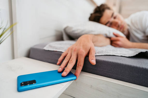 un jeune homme snoozing alarme sur un téléphone intelligent - clock time clock hand urgency photos et images de collection