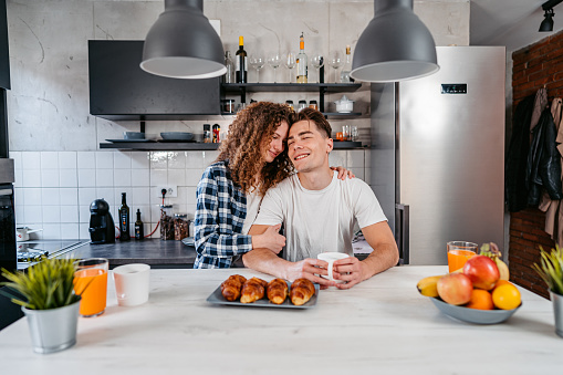 Beautiful young couple standing in the kitchen, having coffee and croissants with juice.