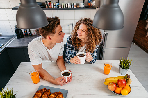 Beautiful young couple standing in the kitchen, having coffee and croissants with juice.