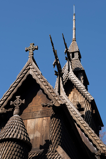 Architectural detail of the roof of Fantoft medieval wooden tarred stave church in the City of Bergen on the west cost of Norway in spring. The image shows details of the roof with dragon heads and a Christian Cross. The dragon heads served to ward off evil spirits and was used by the vikings as to decorate jewellery, accessories and weapons and placed on the bow of their ships (as well as on the roof of buildings). The church was originally built around the year 1150 at Fortun in Sogn (in the Sognefjord). Threatened with demolition as a new church had been built to replace the old, the stave church was bought by consul Fredrik Georg Gade and saved by moving it in pieces to Fantoft in 1883. On 6 June 1992, the church was destroyed by arson. Reconstruction of the church began soon after the fire and was completed in 1997.