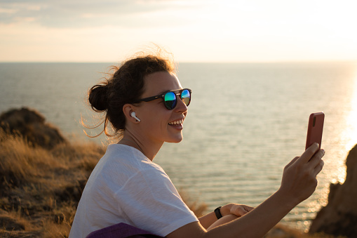 Happy young woman making video call with smartphone, sharing data on social media using 5g internet connection, enjoying sunset scenery view of sea landscape. Travel and adventure lifestyle concept.
