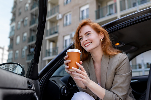 Shot of mid adult woman sitting in her car with open door, contemplating and sipping coffee from a travel mug.