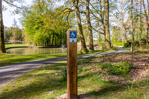 Hiking sign for disabled people in a park, drawing and the word Rolstoel means wheelchair, a path, pond and trees in the background, sunny day in Heidekamp Park, Stein, South Limburg, Netherlands