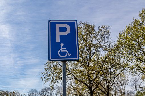Public parking sign for handicap people, blue plate with a P and a drawing of a person in a wheelchair, green foliage and blue sky with few clouds in the background