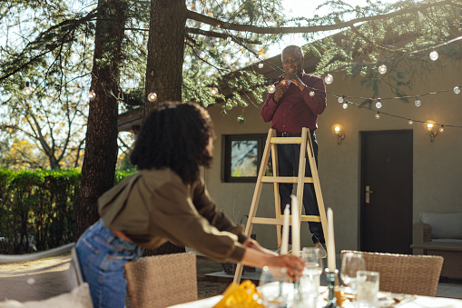 Young african american woman arranging setting on the table for outdoor event dinner, while her father installing electric decoration between trees
