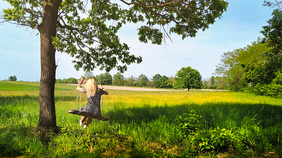 Young woman is enjoying swinging on a tree. Beautiful nature and pastures can be seen in the background.