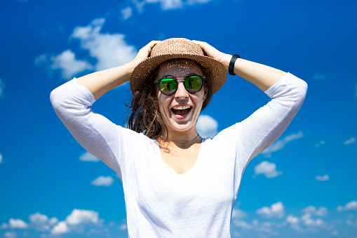 Excited happy young woman in summer hat holding head by two hands opening her mouth in amazement, wearing sunglasses and white t-shirt on blue sky background. Tourist girl astonished receive good news
