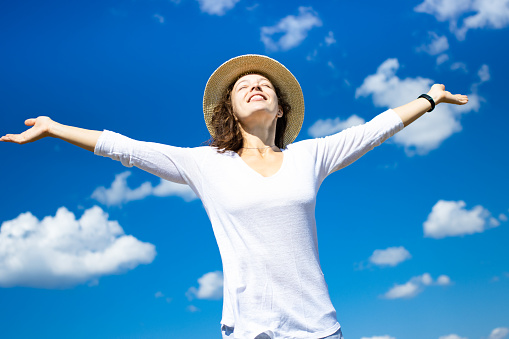 Freedom. Happy young woman in summer hat raising open arms outspread, wearing white cloth on blue sky background. Enjoying life, cheerfulness, happiness, dreams come true lifestyle concept.