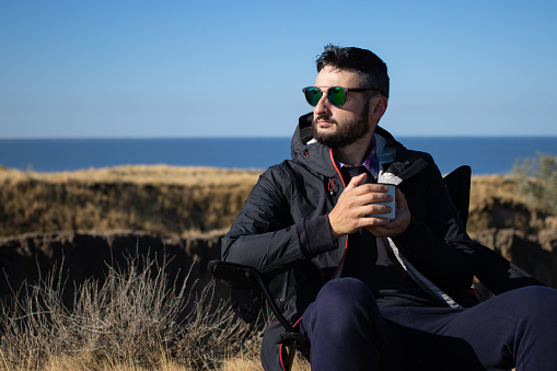 Young brunette man in sunglasses and waterproof jacket holding iron mug cup of coffee, sitting alone on camping chair on cliff by the sea,enjoying vacation in nature landscape.Travel, active lifestyle