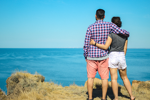 Back view of young couple woman and man traveler enjoying sunny summer scenery view of the sea landscape, hugging each other standing on ocean cliff. Copy space. Travel and adventure lifestyle concept