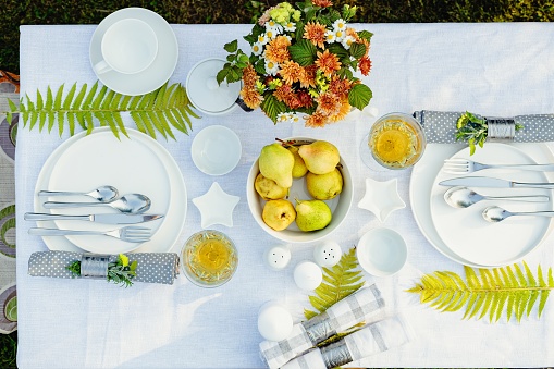 Summer holiday table set up. Elegant table setting with white dishes on a white linen tablecloth. Fruits, drinks in crystal glasses. Table set for outdoor eating in the garden. Top view