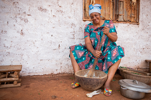 African woman cooks using a mortar and pestle, she is dressed in a traditional dress and she is in the village