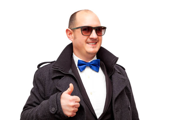 A bald adult man wearing sunglasses and dressed in smart clothes and a blue bow tie is holding up one of his thumbs while smiling. The male is on a white background.