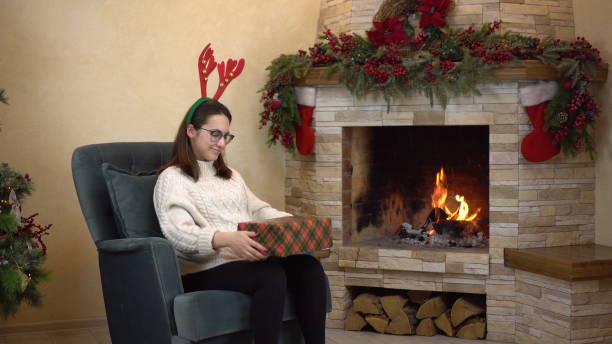 a young pregnant woman with glasses sits in a chair by the fireplace with horns on her head and holds and shakes gifts. christmas mood. - fire match women flame imagens e fotografias de stock